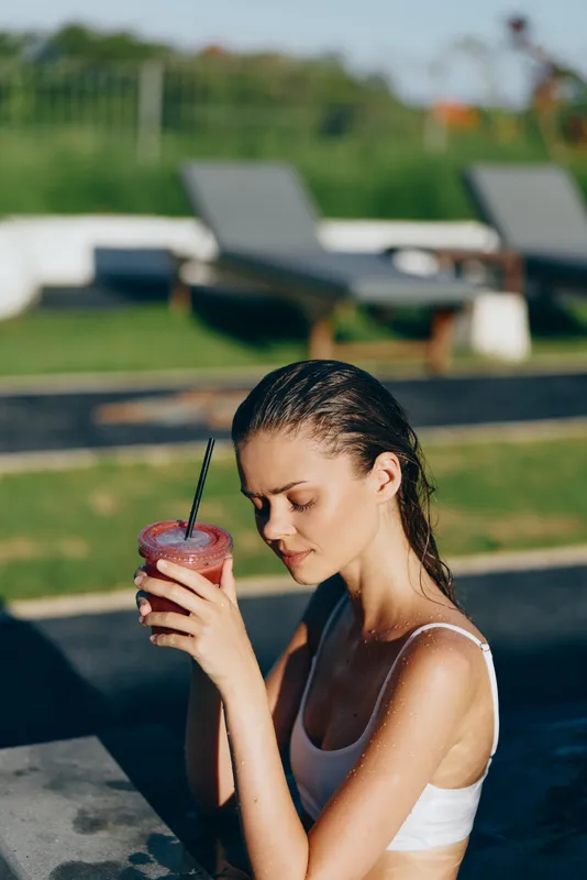 A woman sits by the pool sipping a cold beverage, hair slicked back in a swimsuit. A lifestyle moment of refreshment, leisure, and outdoor summer calm.