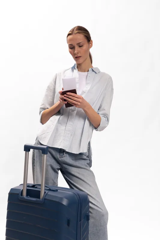 Young woman with suitcase stands and uses smartphone, dressed in casual clothes. Travel concept, modern lifestyle, vacation planning, and communication on the go.