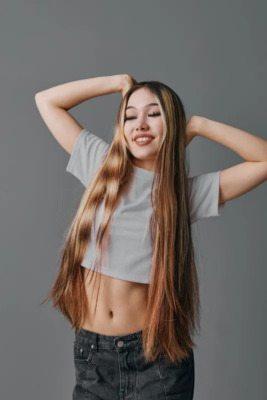 Woman with long straight hair posing with hands behind head wearing casual gray shirt and black jeans against plain background with closed eyes and relaxed expression.
