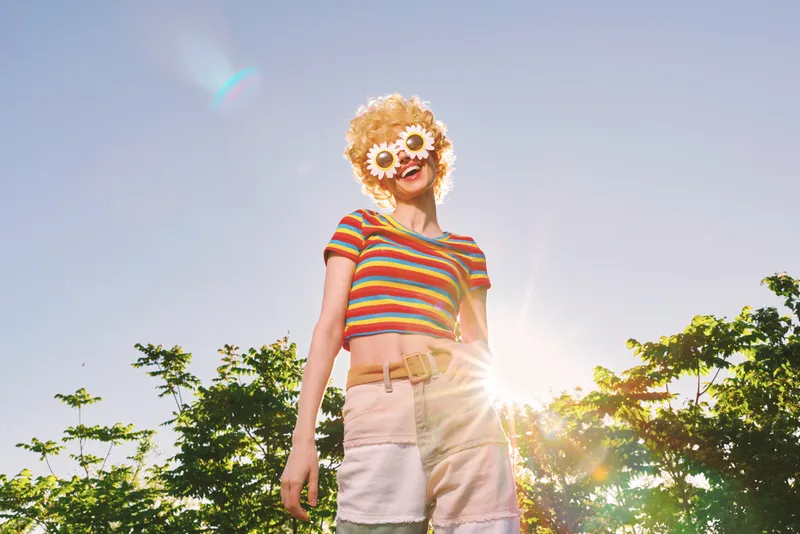 Young woman with a sunflower mask and rainbow striped top stands outdoors under bright sun, creating a playful, whimsical mood and a bold summer vibe against a clear blue sky with lens flare