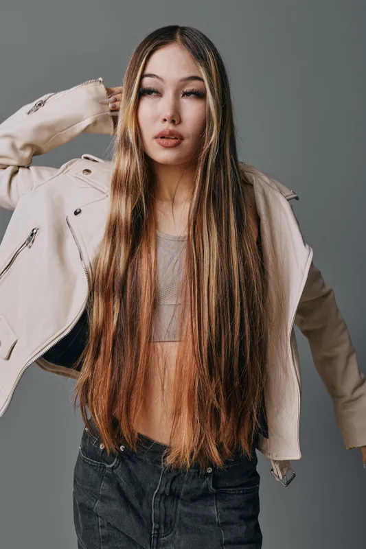 Young woman with long straight hair wearing beige jacket and crop top posing against gray background with confident expression and casual modern fashion style.