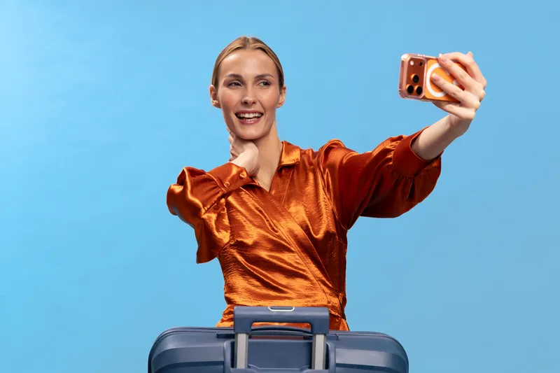 Smiling woman in orange blouse takes selfie with smartphone while standing behind suitcase on blue background. Concept of travel, vacation, tourism, and happy lifestyle.