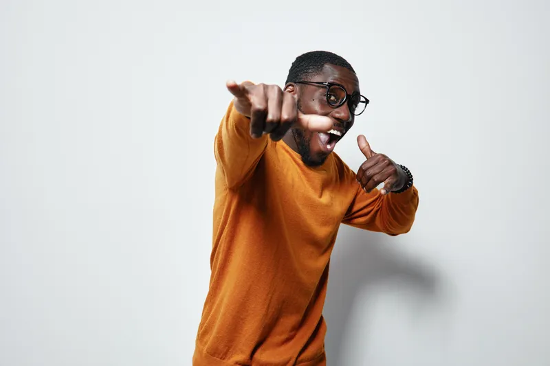 African American man with dark skin wearing glasses and orange sweater pointing and smiling with excitement on white background in studio portrait.