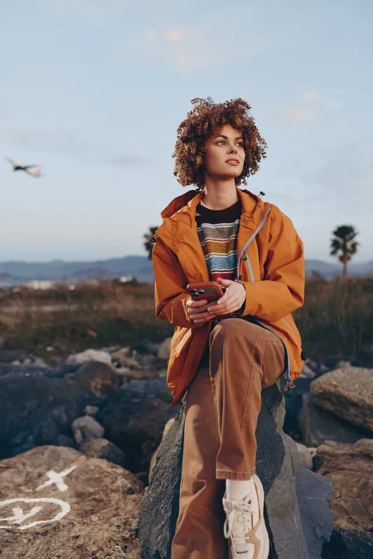 Woman outdoor hiking portrait, curly hair, casual jacket, standing on rocky ground, holding backpack strap, desert landscape behind, soft sunset light, travel lifestyle, relaxed mood