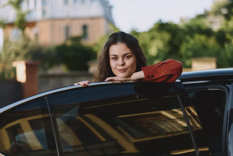A young woman leans on a dark car roof, resting her arms as she gazes softly at the camera in a bright urban setting, conveying a relaxed, everyday moment