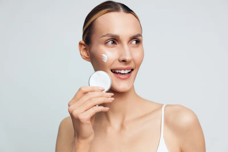 Woman skincare beauty face smile, cotton pad, cream application, young female holds round cosmetic pad near cheek, soft studio light, clean white background, fresh hydrated skin