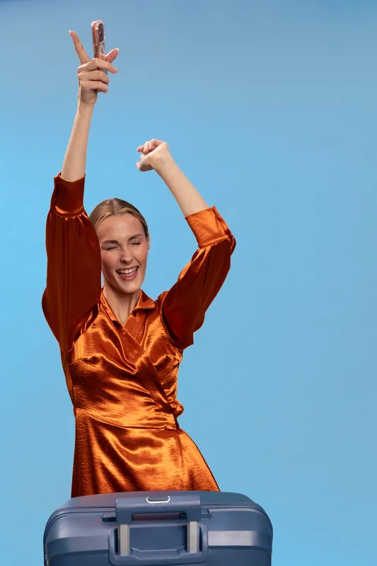 Happy woman in shiny orange dress holds smartphone and dances near blue suitcase on blue background. Concept of travel, vacation, joy, and modern lifestyle.