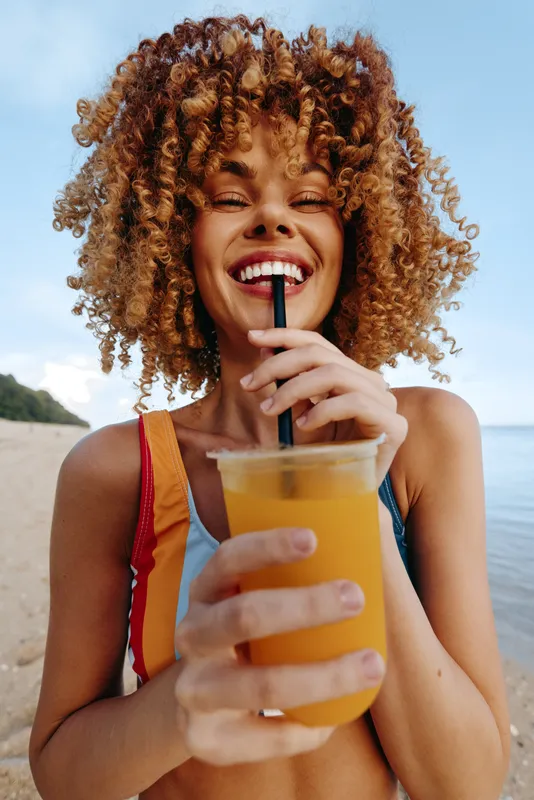 A young woman with curly hair stands on a sunny beach, sipping orange juice from a plastic cup, wearing a striped swimwear top, smiling at the camera with a relaxed, carefree vibe.