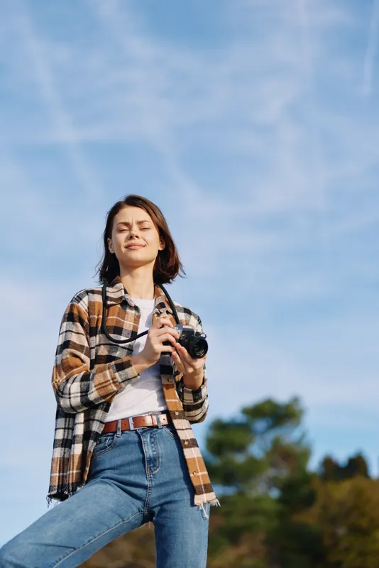 Woman photographer outdoors with a camera, wearing a plaid shirt and jeans, standing under a bright blue sky, capturing a candid moment and natural scenery.