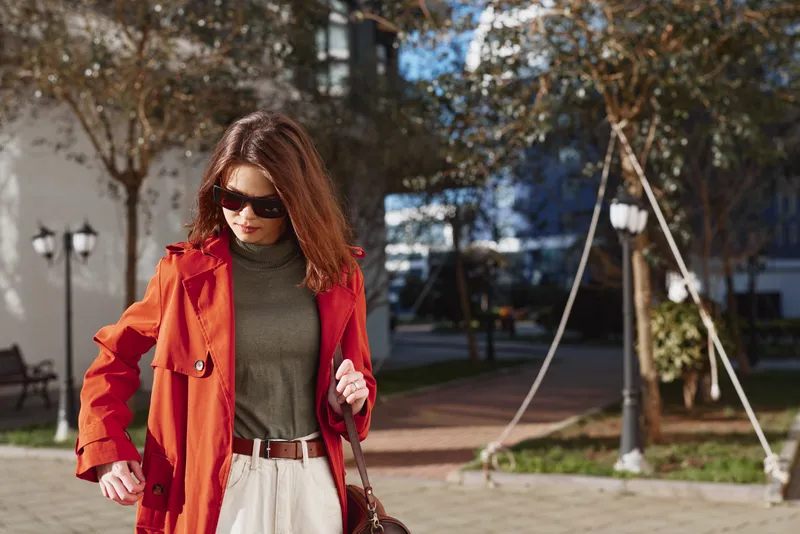 A stylish woman wearing sunglasses and a red coat walks outdoors on a sunny day with trees and urban buildings in the background.