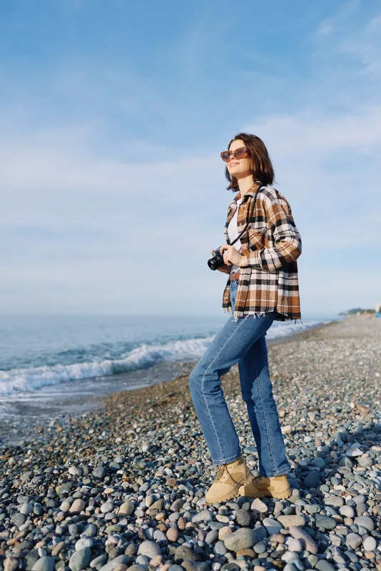 Casual traveler beside the seaside with a camera, wearing a plaid jacket, jeans and boots on a pebble beach. Outdoor photography portrait showcasing exploration and relaxed style against a bright