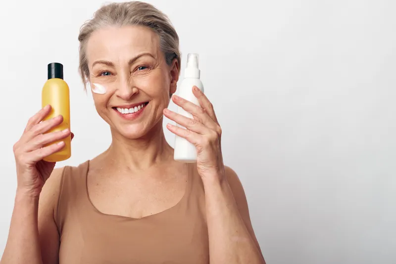 Smiling mature woman with short hair holds two skincare bottles near face with cream applied under eyes. Concept for beauty routine, anti aging, healthy skin and self care at home.