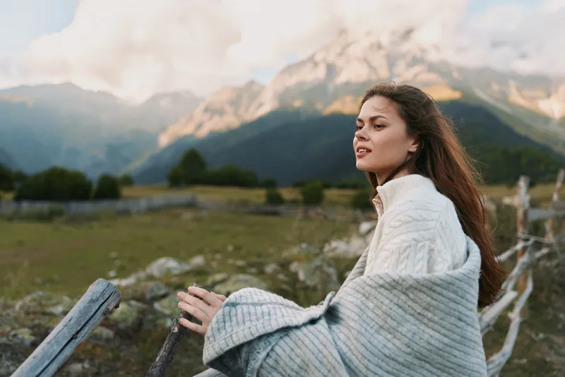 A woman in a white knit sweater sits by a wooden fence, gazing at distant mountains. A calm outdoor lifestyle moment highlighting serenity in nature and horizon.
