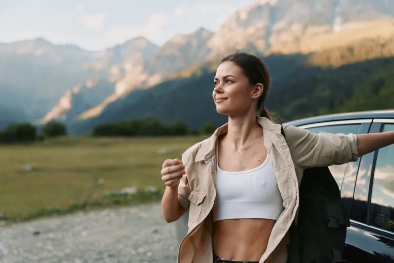 Young woman in a white crop top and beige jacket stands beside a car in a mountain valley, enjoying the fresh air and peaceful nature around her.
