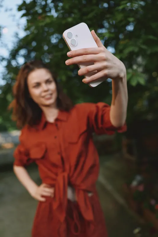 Woman outdoors, wearing a red dress, smiles while taking a selfie in soft sunlight, casual pose, lush greenery, candid lifestyle moment, warm tones, natural mood