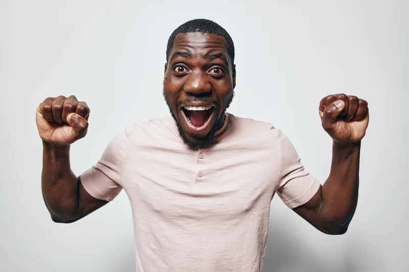 African American man with dark skin wearing light pink shirt celebrating with raised fists and excited facial expression on white background in studio.