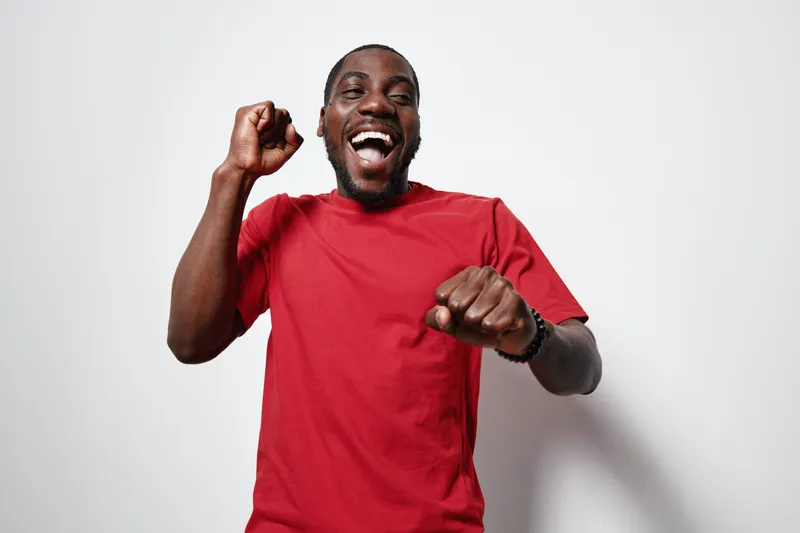 African American man with dark skin wearing red t shirt smiling and celebrating with raised fists on white background. Happy male portrait with joyful expression and casual style.