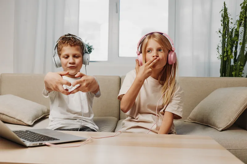 Two children wearing headphones sit on a beige sofa, enjoying music with a laptop nearby. The boy forms a heart shape with his hands while the girl listens thoughtfully, casual home setting.