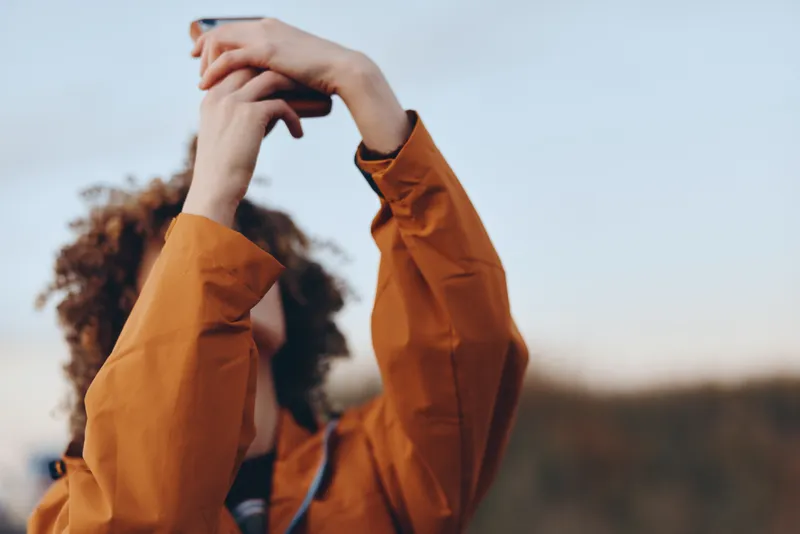 Woman lifestyle outdoors in an orange jacket, curly hair, holding a phone up with both hands for a selfie, capturing nature scenery, soft daylight, blurred field background, close up portrait