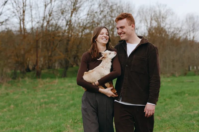 Happy couple outdoors with a small dog, smiling and holding the pet, enjoying a casual park stroll, candid lifestyle moment, warm clothing, autumn backdrop, bright mood, affectionate interaction,