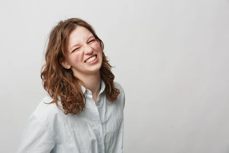 Young woman with curly hair wearing white shirt smiles with eyes closed against plain background. Natural expression and casual style create relaxed and joyful mood in studio setting.
