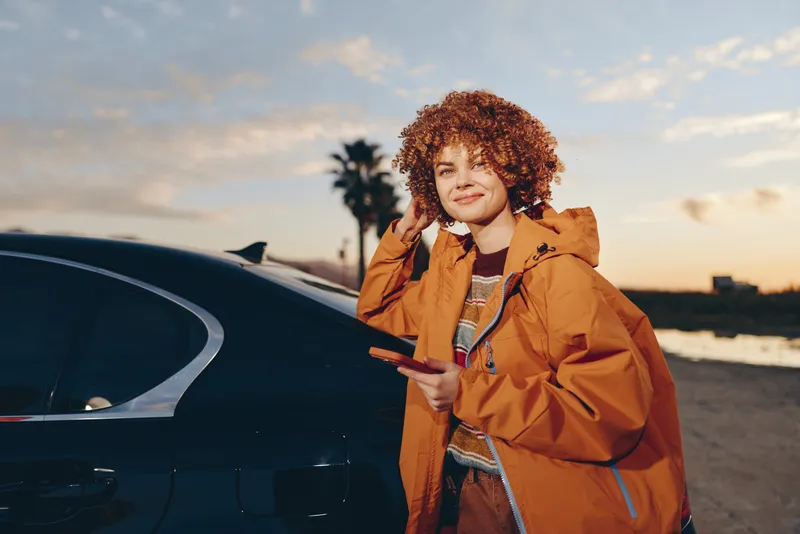 Woman lifestyle outdoor, standing by car at sunset, smiling, wearing jacket, relaxed travel mood, coastal road background, golden sky, soft breeze, warm evening light