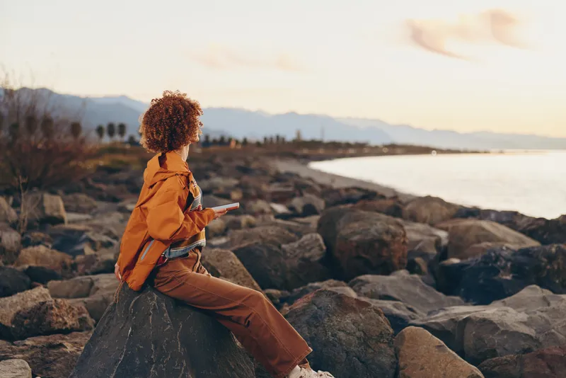 Woman with curly hair sitting on rocks, facing the ocean at sunset, outdoors lifestyle travel adventure, scenic coast, warm light, calm waves, peaceful evening, nature