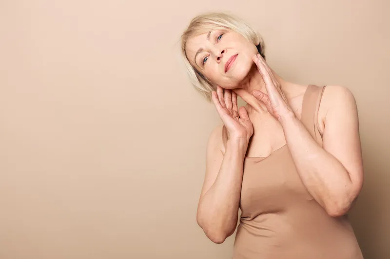 Elegant mature woman with short blonde hair gently touches her neck and face, wearing a beige tank top. Neutral background highlights skin care, beauty, and aging gracefully concepts.