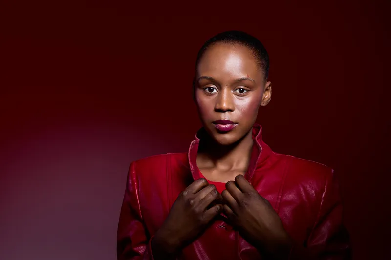 Portrait of confident black woman with dark skin wearing red leather jacket, posing against dark red background. Concept of fashion, beauty, and empowerment in studio lighting.