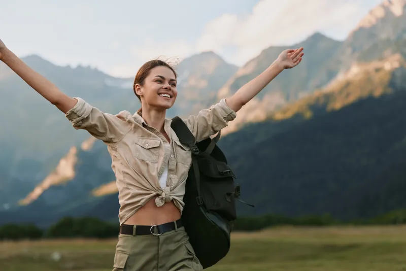 A joyful woman hiking outdoors with a backpack, arms raised in celebration. The expansive nature and distant mountains frame a moment of adventure and freedom. This lifestyle scene captures