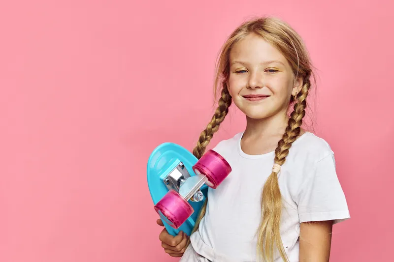 Smiling girl with long braided hair holding blue skateboard against pink background. Casual outfit with white t-shirt. Youth lifestyle concept.