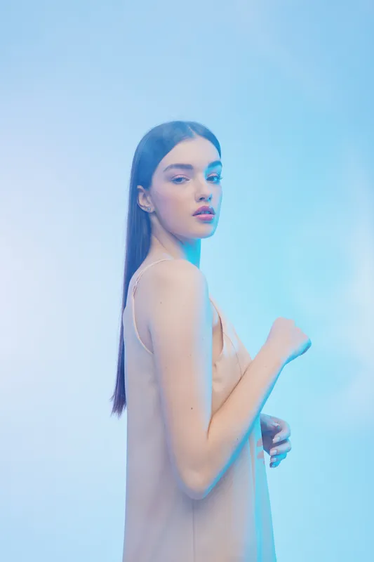 Young woman with long dark hair poses in beige dress against blue background. Concept of beauty, fashion, elegance, and modern style with soft lighting effect.