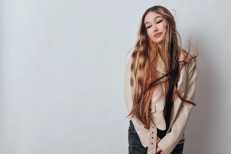 Young woman with long hair wearing light jacket and jeans posing against plain wall with closed eyes and relaxed expression in studio portrait for fashion concept.