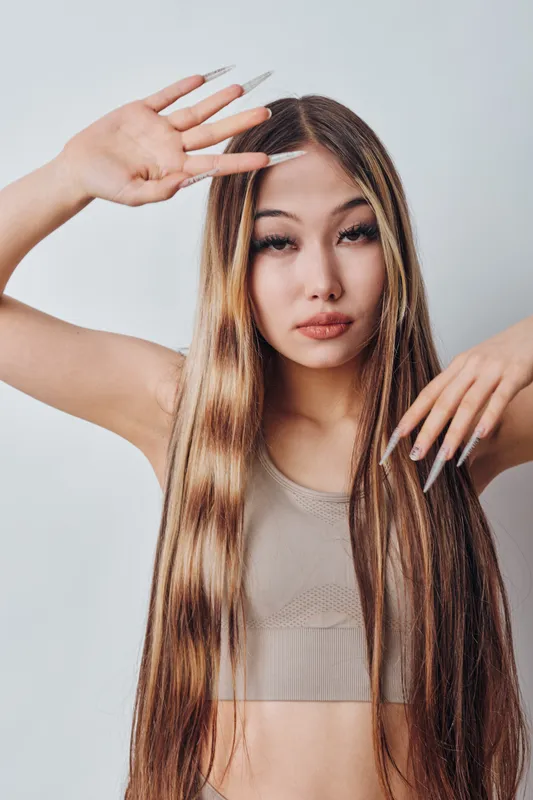 Young woman with long straight hair and natural makeup posing with hands near face in studio. Casual clothing and smooth skin with neutral expression on light background.