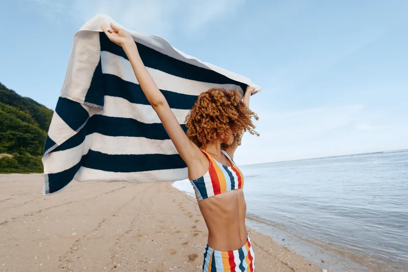 A woman on a sunny beach lifts a striped towel above her head, wearing a colorful swimsuit. A bright, carefree moment for travel, leisure, and summer joy in motion.