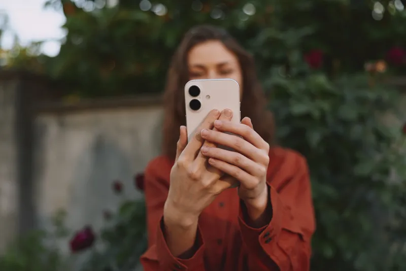 A young woman takes a candid selfie outdoors with a smartphone, smiling softly as she holds the device up in a casual garden setting