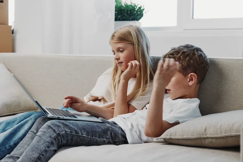 Two children sitting on a beige sofa using a laptop together in a bright living room. Concept of learning, technology, family, childhood, and casual home environment.