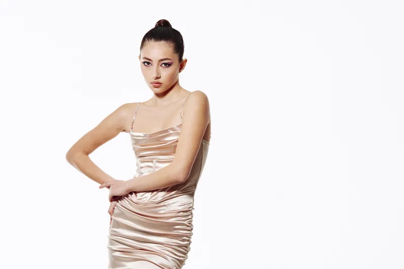 Fashion model in a silky beige dress poses elegantly against white background with sleek hairstyle and sharp makeup, embodying beauty and creative fashion concepts.