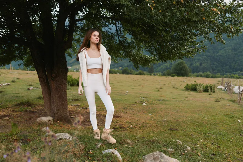 Young woman in a white outfit stands in a green field under a large tree with a relaxed, confident expression. Her hands rest at her sides as soft natural light filters through leaves, creating a