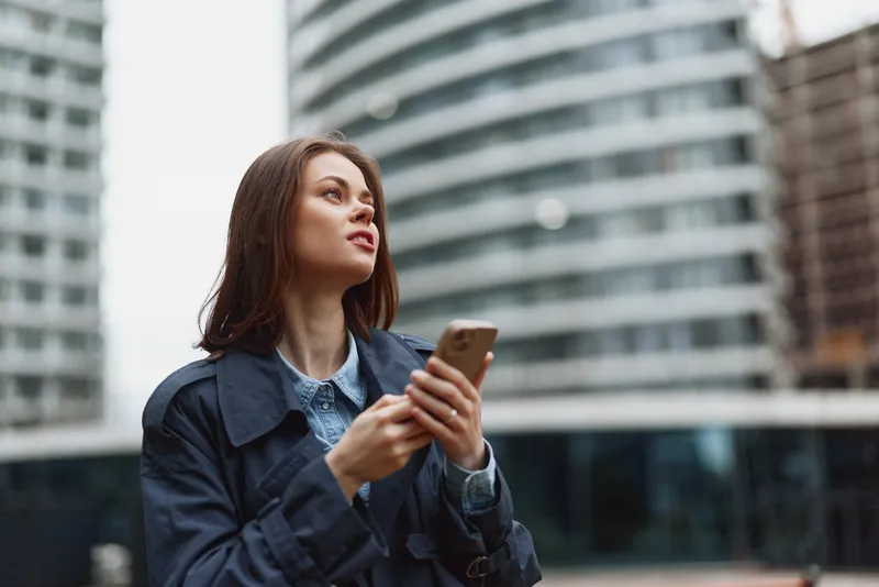 Young woman holding smartphone looking up thoughtful expression casual clothing modern city background daytime urban environment