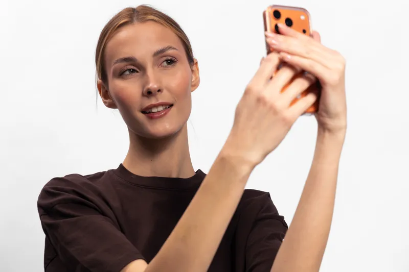 Young woman in brown shirt takes a selfie with smartphone. Studio portrait on white background shows casual style and natural smile. Concept for social media and communication.
