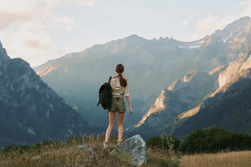Young woman with backpack stands on a rock overlooking distant mountains, feeling adventurous, free, hopeful, and connected to the outdoors in bright daylight