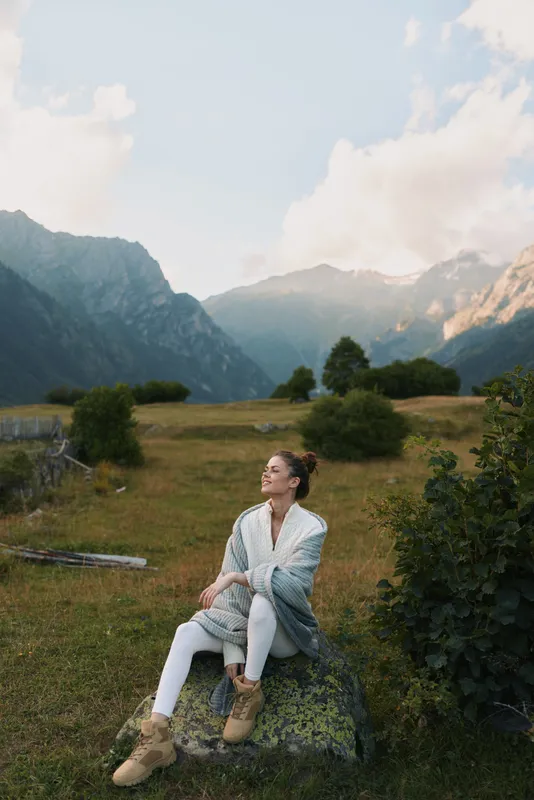 A woman in casual clothes sits on a rock wrapped in a blanket enjoying the mountain landscape during daytime. Concept of lifestyle, relaxation, and nature connection.