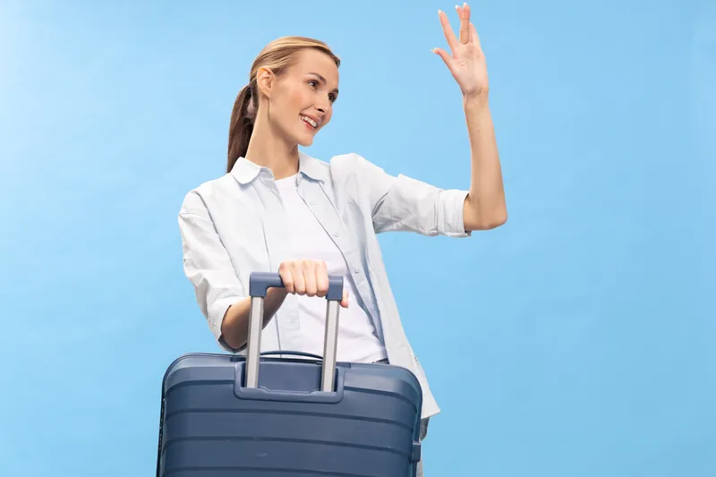 Smiling woman in casual white shirt holds navy blue suitcase handle and waves hand isolated on light blue background. Concept of travel, vacation, tourism, and happy departure.