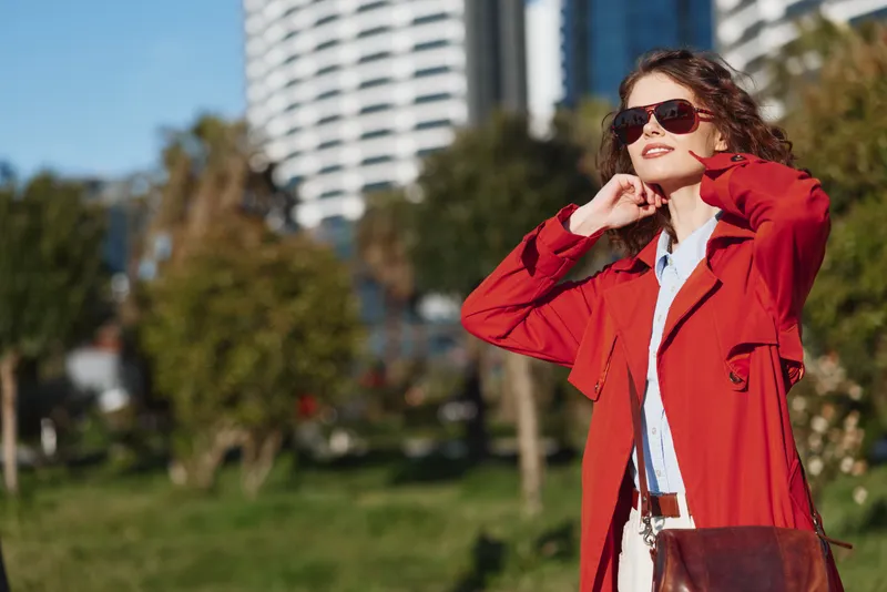 Young woman wearing a red coat stands outdoors in a sunny city park, adjusting her collar with a confident smile as the scene feels relaxed and fashion forward.