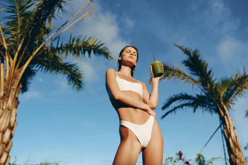 A confident woman in a white bikini stands outdoors under a clear blue sky, holding a green smoothie. Lifestyle shot highlighting sunshine, leisure, and summer relaxation, with palm trees framing the