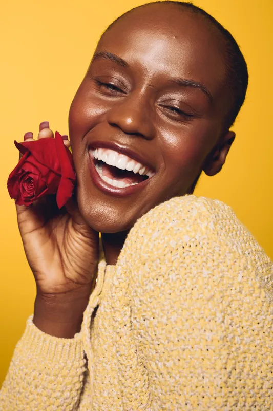 Happy smiling black woman with dark skin holding red rose near face on yellow background. Close-up portrait of joyful African female model in cozy beige sweater. Concept of beauty and happiness.