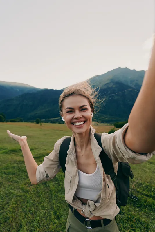 A smiling young woman outdoors raises her arms in a joyful gesture while exploring a grassy valley with distant mountains and a bright, clear sky.