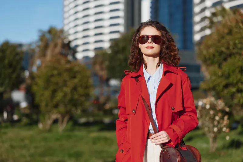 Young woman in a red coat stands confidently on a sunny city street with sunglasses, modern buildings in the background, her expression composed and stylish in natural daylight.