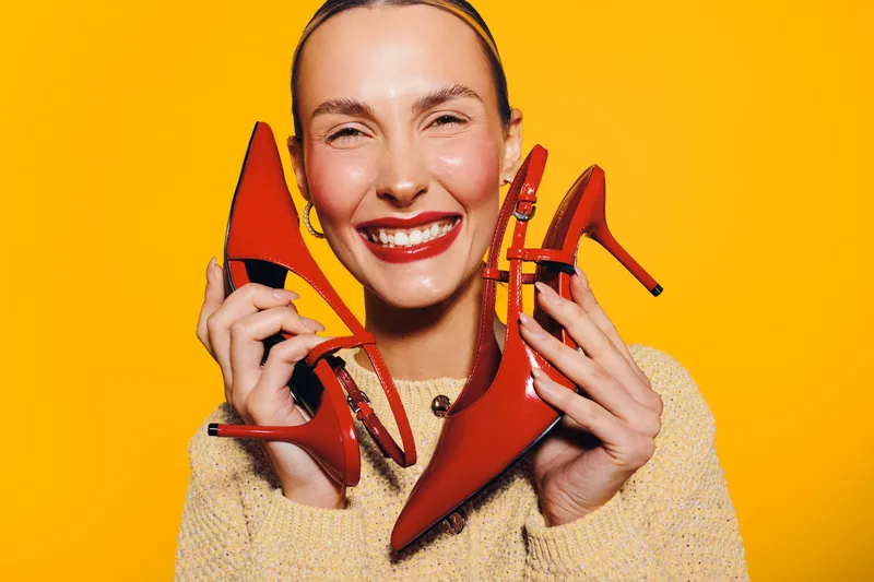 Smiling woman holds red high heel shoes near face on bright yellow background. Concept of fashion, style, footwear, and joyful emotions in studio portrait with vibrant colors.